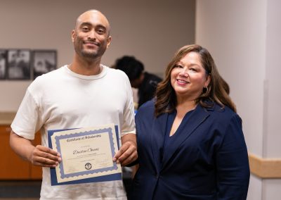 A person in a white t-shirt holds a scholarship certificate while standing next to another person in a navy blazer, both smiling in an indoor setting.