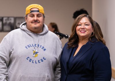 Two individuals pose together at an event, one wearing a Fullerton College hoodie and a yellow cap, the other in a dark blazer.