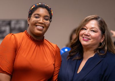 Two individuals smile warmly at the camera, one wearing an orange top with a colorful headband and glasses, the other in a navy blazer.