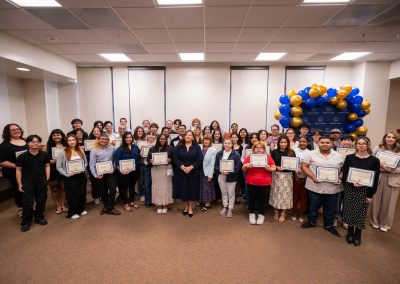 A group of individuals stands together in a room, holding certificates, with a blue and gold balloon arch in the background.