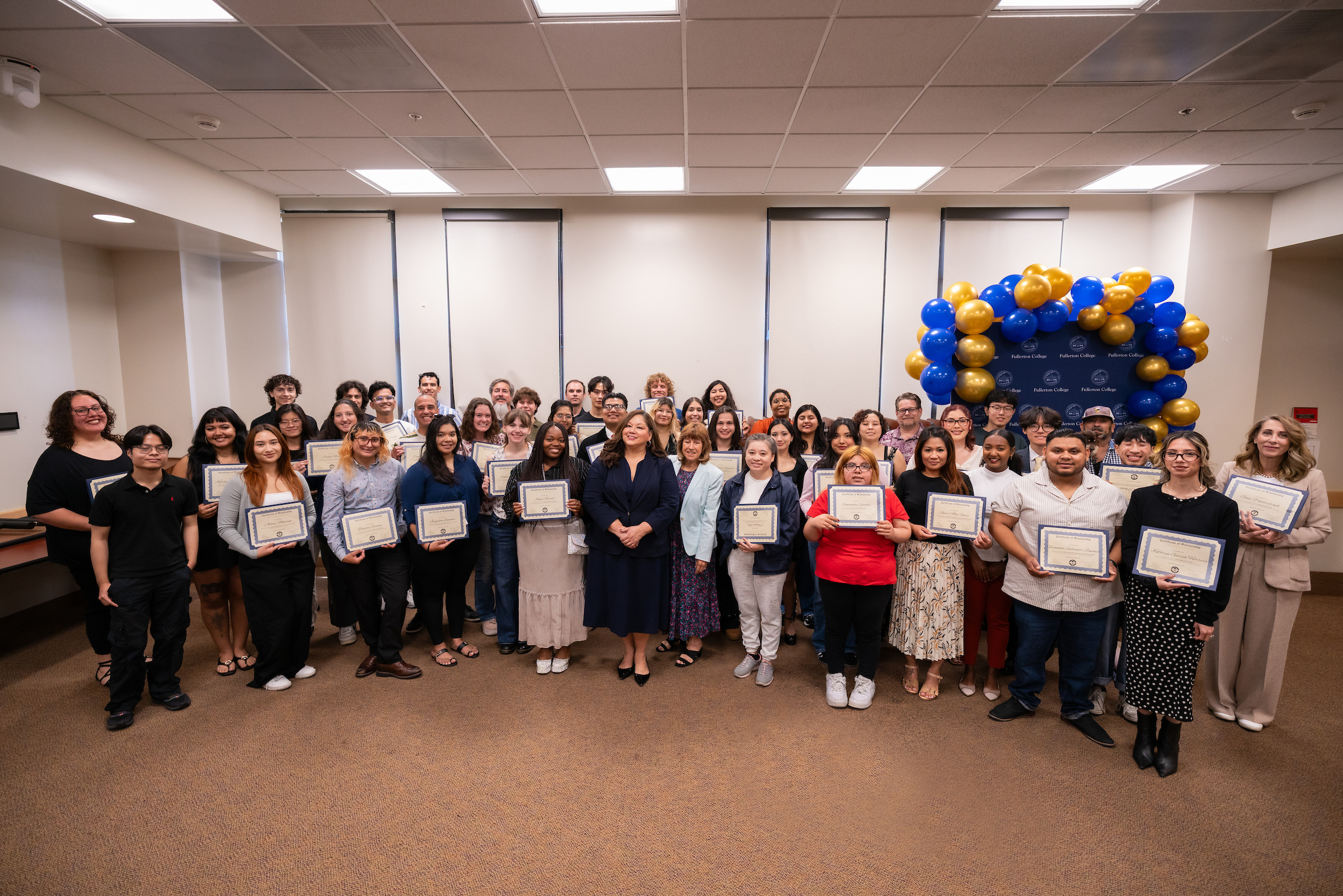A group of individuals stands together in a room, holding certificates, with a blue and gold balloon arch in the background.