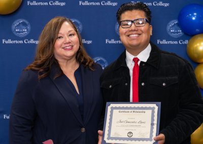 A person in a black jacket and red tie holds a scholarship certificate while standing next to another person in a navy blazer, both smiling in front of a blue backdrop with “Fullerton College” branding and balloons.