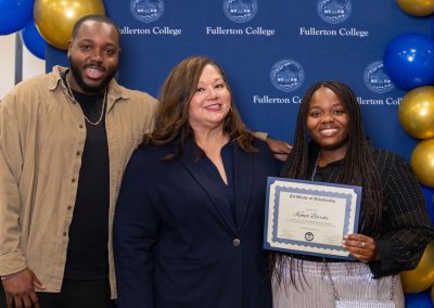 Three individuals pose together at a Fullerton College event, with one holding a scholarship certificate, against a backdrop of blue and gold balloons.