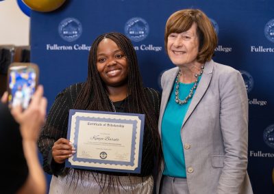 A young person holds a scholarship certificate while standing next to an older individual at a Fullerton College event, with someone capturing the moment on a smartphone.