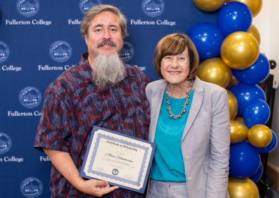 Two individuals stand together in front of a blue backdrop with "Fullerton College" branding and blue and gold balloons, one holding a certificate of scholarship.