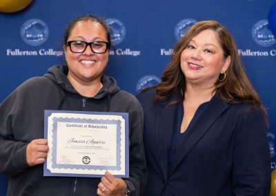 A person in a hoodie holds a scholarship certificate while standing beside another person in a blazer, both smiling in front of a blue backdrop with “Fullerton College” branding.