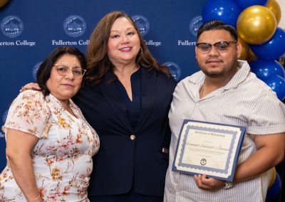 Three individuals pose together at a Fullerton College event, with one holding a scholarship certificate, against a backdrop of balloons and the college logo.