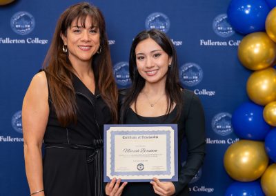 Two individuals stand together in front of a blue backdrop with "Fullerton College" branding, holding a certificate of scholarship.