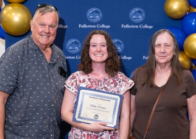 A young person holds a scholarship certificate while standing with two adults in front of a backdrop featuring the name “Fullerton College” and balloons.