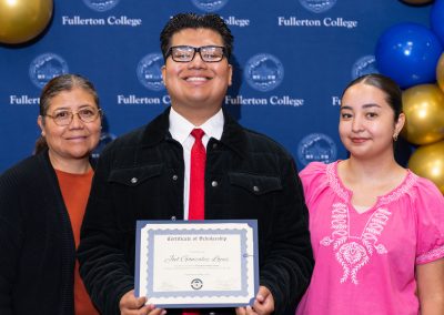 A young person holds a scholarship certificate while standing with two adults in front of a backdrop featuring the Fullerton College logo and balloons.