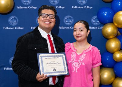 Two individuals stand together in front of a blue backdrop with "Fullerton College" branding, one holding a scholarship certificate, surrounded by blue and gold balloons.