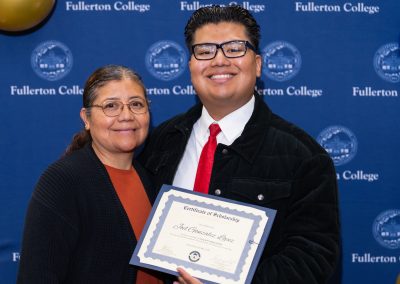 A young person holds a scholarship certificate while standing with an adult beside them, both smiling in front of a blue backdrop featuring the Fullerton College logo.